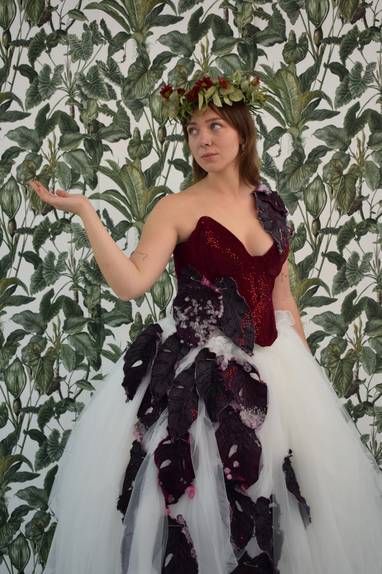 Wilting Grace Frau in einzigartigem Kleid mit Blumenkranz, vor einer Pflanzenmuster-Wand.